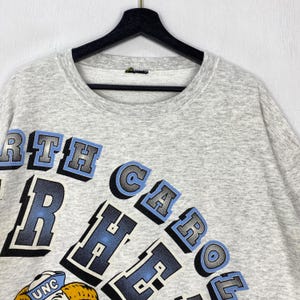 May include: A light grey heathered crewneck sweatshirt featuring the text NORTH CAROLINA in bold, blocky blue and black letters with a drop shadow effect. A partial graphic of a mascot wearing a blue hat with the letters UNC is visible below.