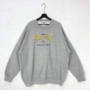 Può includere: Una felpa girocollo grigio melange con la scritta Nashville Predators ricamata in giallo e blu navy sul petto. L'etichetta del marchio Lee è visibile all'interno del colletto. Il capo presenta maniche lunghe e polsini a costine.