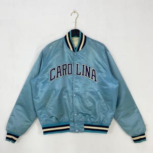 May include: Light blue satin bomber jacket with the word "CAROLINA" across the chest in navy blue and white lettering. The jacket has a striped collar, cuffs, and hem in navy, white, and light blue.