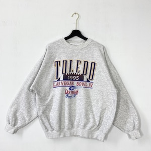 Könnte beinhalten: Ein graues Sweatshirt mit dem Wort "TOLEDO" in Marineblau und goldfarbenen Buchstaben. Darunter steht der Text "1995 LAS VEGAS BOWL IV" in Rot und Blau. Eine Football-Grafik befindet sich in der Mitte des Designs.