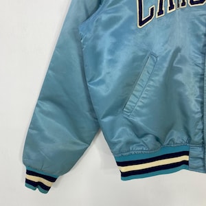 May include: Light blue satin jacket with a ribbed collar and cuffs. The cuffs feature navy blue, white, and light blue stripes. The jacket has a pocket and the letters "LA" in white on the upper right side.