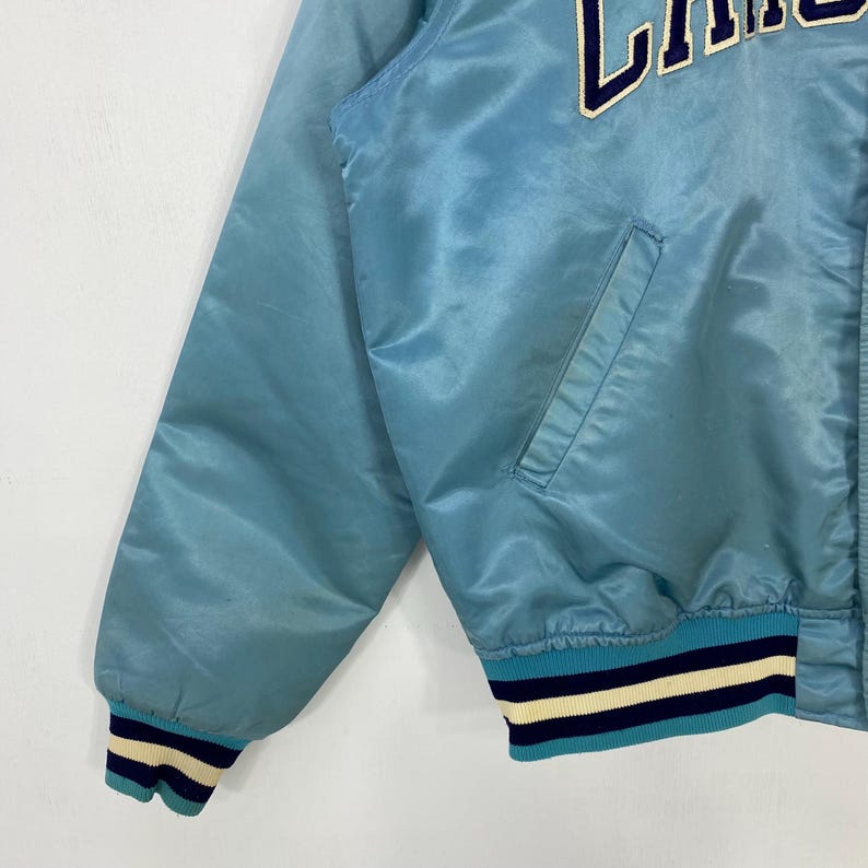 May include: Light blue satin jacket with a ribbed collar and cuffs. The cuffs feature navy blue, white, and light blue stripes. The jacket has a pocket and the letters "LA" in white on the upper right side.