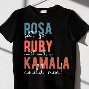 May include: Black t-shirt with a graphic print that reads "ROSA sat, so RUBY could walk, so KAMALA could run!"