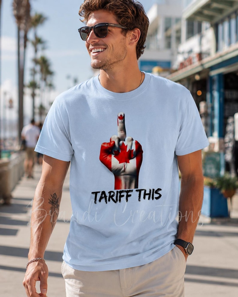 Puede incluir: Una camiseta azul claro con un gr&aacute;fico de una mano pintada con el dise&ntilde;o de la bandera canadiense, mostrando el dedo medio. Debajo del gr&aacute;fico, el texto TARIFF THIS est&aacute; impreso en una fuente negra, negrita y con efecto desgastado.