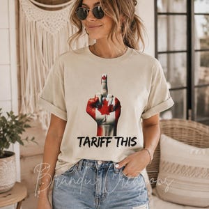 Puede incluir: Una camiseta beige de manga corta con un gr&aacute;fico de una mano pintada con el dise&ntilde;o de la bandera canadiense, mostrando un gesto de dedo medio. Debajo del gr&aacute;fico, el texto TARIFF THIS est&aacute; impreso en una fuente negra, negrita y desgastada.