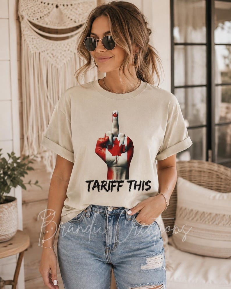 Puede incluir: Una camiseta beige de manga corta con un gr&aacute;fico de una mano pintada con el dise&ntilde;o de la bandera canadiense, mostrando un gesto de dedo medio. Debajo del gr&aacute;fico, el texto TARIFF THIS est&aacute; impreso en una fuente negra, negrita y desgastada.