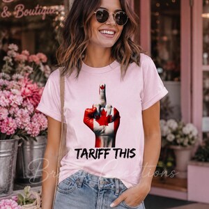 Puede incluir: Una camiseta de manga corta de color rosa claro con un gr&aacute;fico de una mano pintada con el dise&ntilde;o de la bandera canadiense, mostrando el dedo coraz&oacute;n. Debajo del gr&aacute;fico, el texto negro dice TARIFF THIS en una fuente negrita y desgastada.