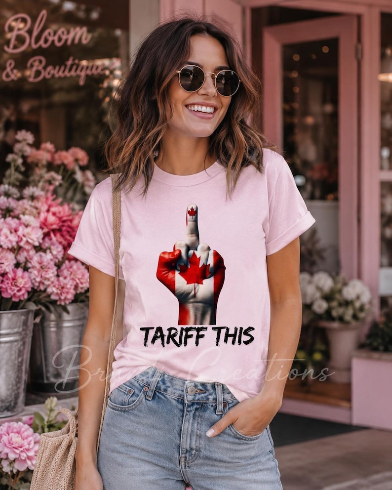 Puede incluir: Una camiseta de manga corta de color rosa claro con un gr&aacute;fico de una mano pintada con el dise&ntilde;o de la bandera canadiense, mostrando el dedo coraz&oacute;n. Debajo del gr&aacute;fico, el texto negro dice TARIFF THIS en una fuente negrita y desgastada.