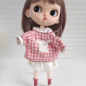 May include: A doll wearing a pink and white checkered shirt with a white bear applique, white shorts, red socks, and white sneakers.