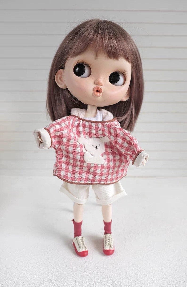 May include: A doll wearing a pink and white checkered shirt with a white bear applique, white shorts, red socks, and white sneakers.
