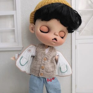 May include: A doll with black hair wearing a brown knitted vest, a white shirt, blue jeans with red embroidery that says "Hug Me", and a white jacket with green trim. The doll is wearing a yellow knitted hat.