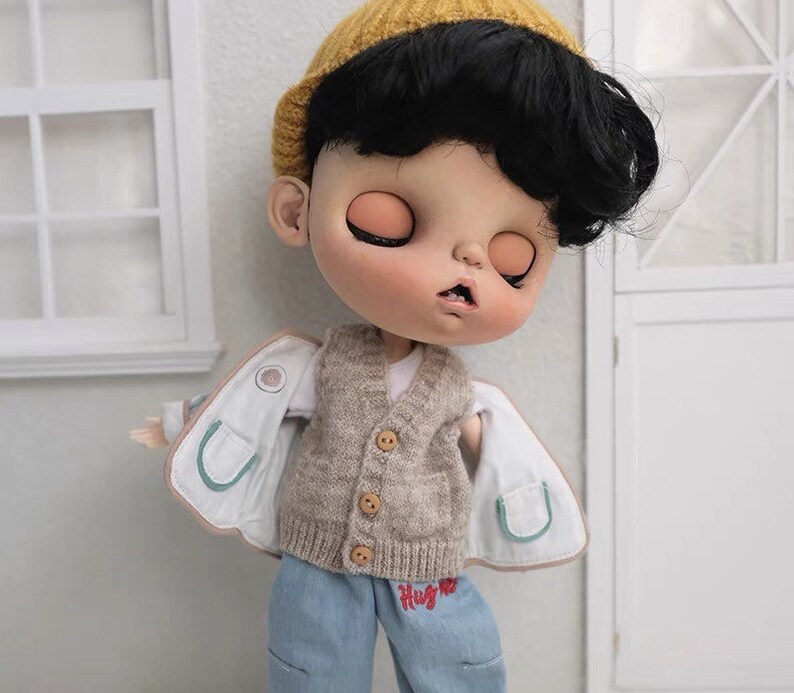May include: A doll with black hair wearing a brown knitted vest, a white shirt, blue jeans with red embroidery that says "Hug Me", and a white jacket with green trim. The doll is wearing a yellow knitted hat.