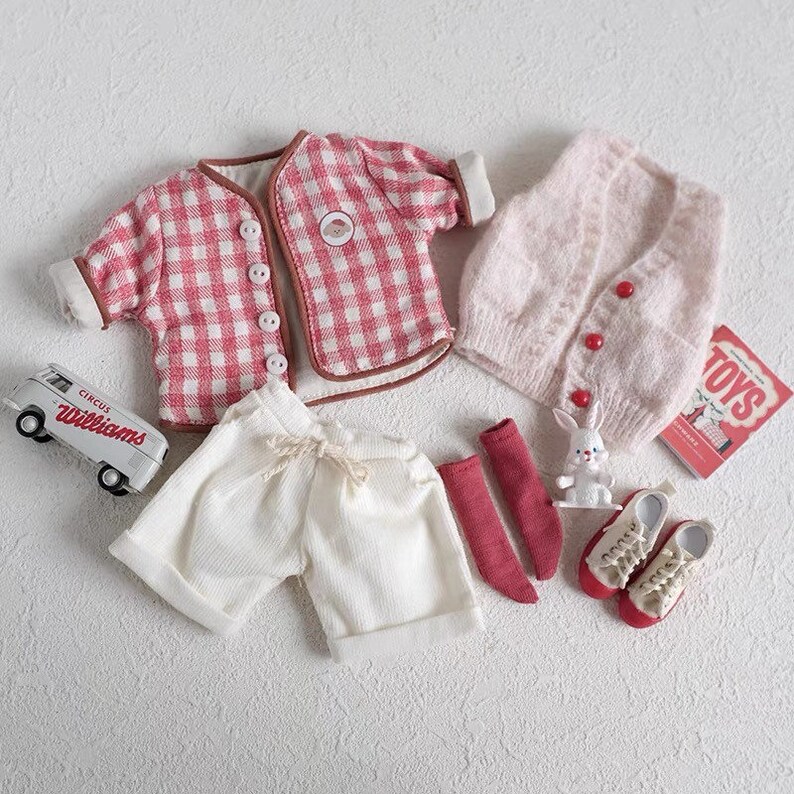 May include: A doll outfit set with a red and white gingham shirt, a pink knitted vest, white shorts, red socks, and white sneakers. The set also includes a toy van, a toy bunny, and a book titled "TOYS".