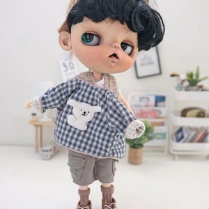 May include: A doll wearing a blue and white plaid shirt with a white bear applique, brown cargo shorts, brown socks, and brown sneakers. The doll has black hair, green eyes, and is wearing a brown knitted hat.