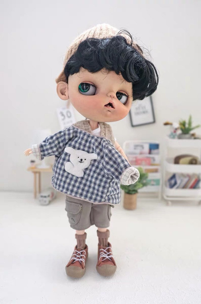 May include: A doll wearing a blue and white plaid shirt with a white bear applique, brown cargo shorts, brown socks, and brown sneakers. The doll has black hair, green eyes, and is wearing a brown knitted hat.