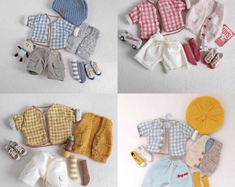 May include: Four different doll outfits for a baby doll. Each outfit includes a shirt, pants, socks, and a cardigan. The shirts are all plaid and the pants are all white. The cardigans are in different colors and styles.