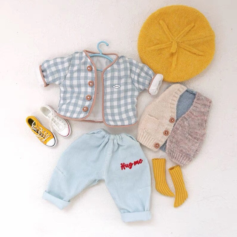 May include: A baby outfit set with a blue and white gingham jacket, a light blue denim pants with "Hug me" embroidered in red, a beige and gray knitted vest, a yellow beret, and yellow socks.