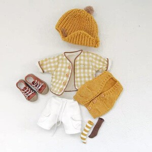 May include: A yellow knitted hat with a pom-pom, a white and yellow plaid jacket, a yellow knitted vest, white shorts, brown and white striped socks, and brown shoes.