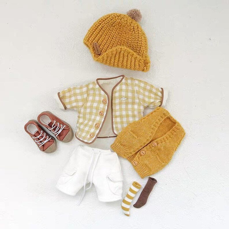 May include: A yellow knitted hat with a pom-pom, a white and yellow plaid jacket, a yellow knitted vest, white shorts, brown and white striped socks, and brown shoes.