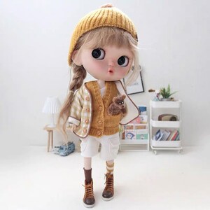 May include: A doll wearing a yellow and white plaid jacket, a yellow knitted cardigan with a brown teddy bear sewn on the front, white shorts, yellow and white striped socks, and brown boots. The doll has blonde hair in a braid and is wearing a yellow knitted hat.