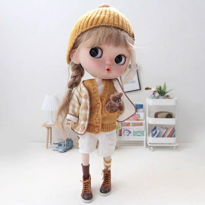 May include: A doll wearing a yellow and white plaid jacket, a yellow knitted cardigan with a brown teddy bear sewn on the front, white shorts, yellow and white striped socks, and brown boots. The doll has blonde hair in a braid and is wearing a yellow knitted hat.