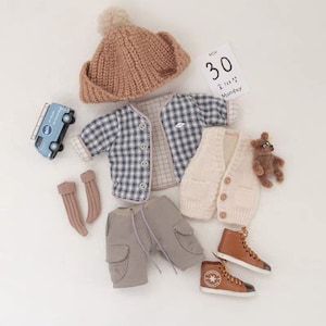 May include: A baby outfit set with a brown knit hat, a blue and white plaid shirt, a cream knit vest, gray cargo shorts, brown leather high-top sneakers, and brown knit socks. The set also includes a small blue toy van and a teddy bear.
