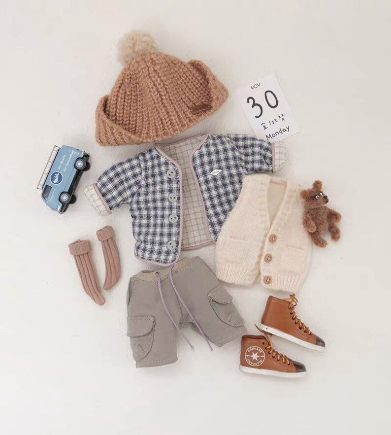 May include: A baby outfit set with a brown knit hat, a blue and white plaid shirt, a cream knit vest, gray cargo shorts, brown leather high-top sneakers, and brown knit socks. The set also includes a small blue toy van and a teddy bear.