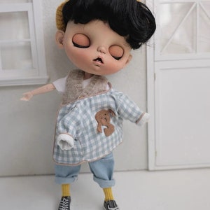 May include: A doll wearing a blue and white plaid shirt with a brown bear patch, yellow socks, and black sneakers. The doll has black hair and is wearing a yellow knitted hat.