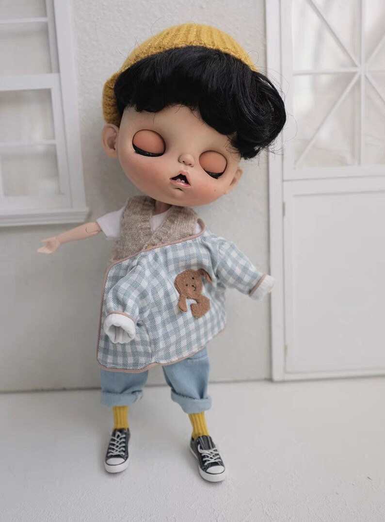 May include: A doll wearing a blue and white plaid shirt with a brown bear patch, yellow socks, and black sneakers. The doll has black hair and is wearing a yellow knitted hat.
