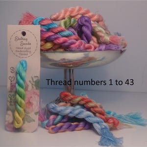 May include: A collection of 43 hand-dyed embroidery threads in various shades of pink, blue, green, and purple. The threads are displayed in a silver bowl and on a white surface. The label reads "Shifting Sands Hand dyed Embroidery Thread" and "69 Skating Sand".