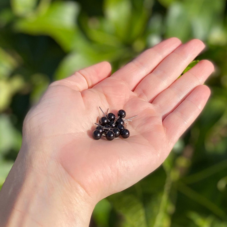 May include: A hand holding a group of black glass beads with silver pins. The beads are arranged in a cluster and are all the same size.