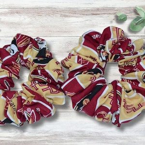 May include: Two scrunchies in maroon, gold, and black fabric. The scrunchies are made of a patterned fabric with the letters "FSU" and other graphics. The scrunchies are on a light wood-grain surface.