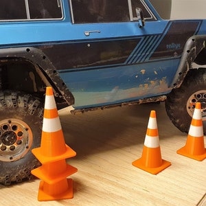 May include: A blue toy truck with black tires and a black and white stripe design on the side. The truck is surrounded by four orange traffic cones.