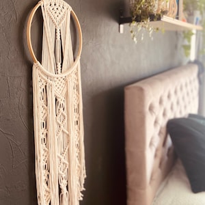 以下が含まれることがあります： 木製の輪とフリンジが付いた白いマクラメの壁掛け。壁掛けは灰色の壁と白いヘッドボードの前にある。