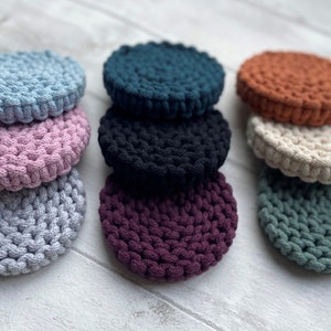 Puede incluir: Un conjunto de diez posavasos redondos de crochet en varios colores, incluyendo azul claro, rosa, gris, negro, verde azulado, burdeos y beige. Los posavasos están apilados en pares y dispuestos sobre una superficie de madera blanca.