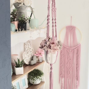 Pode incluir: Um suporte de plantas em macramê rosa com um vaso de cerâmica branco e flores cor-de-rosa penduradas nele. O suporte está na frente de uma prateleira branca com uma variedade de plantas e decoração.