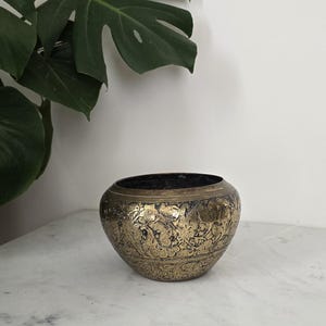 May include: A decorative, antique-style brass bowl with intricate floral patterns. The bowl is round with a dark interior and sits on a marble surface. A large green leaf is visible in the background.