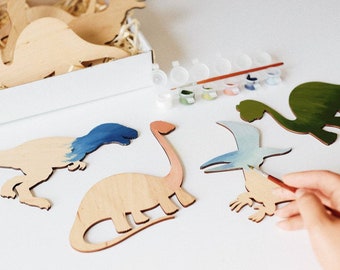 May include: A set of wooden dinosaur shapes for painting. The dinosaurs are in various poses, including a T-Rex, a long-necked dinosaur, and a flying dinosaur. The set includes a small container of paint and a paintbrush.