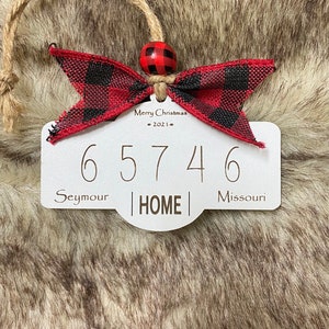 May include: A white wooden Christmas ornament with the numbers 65746, the word HOME, and the text "Seymour Missouri". It has a red and black plaid bow, a red and black bead, and a jute string for hanging. The text "Merry Christmas - 2021" is also present.