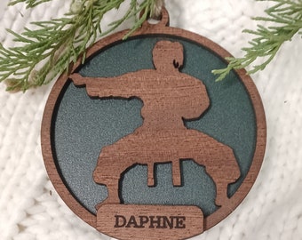 May include: A circular wooden ornament depicting a karate figure in a fighting pose. The figure is brown, set against a dark green backdrop. The name "DAPHNE" is displayed on a small rectangular plaque. A jute string is attached for hanging.