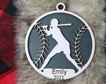 May include: A wooden baseball ornament with a white batter silhouette on a dark teal background. The ornament has a baseball design with white stitching and the name "Emily" and the year "2021" engraved on it. The ornament has a hole for hanging.