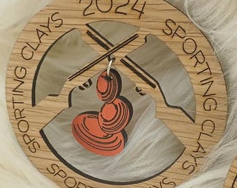 May include: Wooden ornament with the text "2024" and "Sporting Clays" engraved around the edge. The ornament features a design of crossed shotguns and orange clay targets. A red bead and twine are attached for hanging.