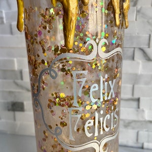 May include: A clear tumbler with gold glitter and a gold drip design. The tumbler has the words "Felix Felicis" written in silver lettering.