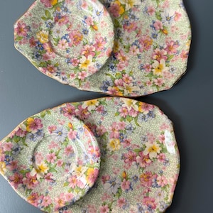 Może przedstawiać: Dwa zestawy talerzy w kwiaty w stylu vintage. Każdy zestaw zawiera duży, owalny talerz i mniejszy, okrągły talerz. Talerze mają jasnozielone tło z kolorowym kwiatowym wzorem w kolorze różowym, żółtym i niebieskim. Krawędzie są karbowane i złote.