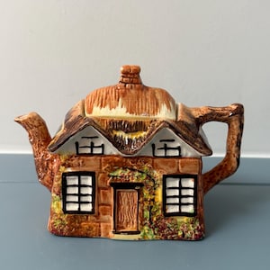 May include: A ceramic teapot shaped like a cottage with a thatched roof, brown brick walls, and black-framed windows. The spout and handle resemble tree branches, and the front features a central door surrounded by painted green and red foliage.