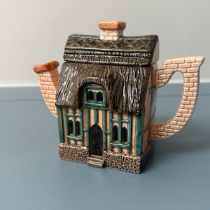 Puede incluir: Una tetera de cerámica con forma de cabaña con techo de paja, un pico con patrón de ladrillos y un asa. La estructura presenta entramado de madera verde, una puerta arqueada oscura, varias ventanas y una base de piedra texturizada en tonos marrón y beige.