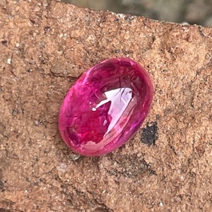 May include: A vibrant pink oval-shaped gemstone with a smooth, polished surface. The gemstone is set against a rough, brown background.