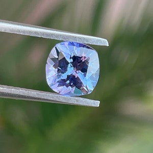 May include: A cushion-cut, purple gemstone with a light blue hue, held by tweezers against a blurred green background.