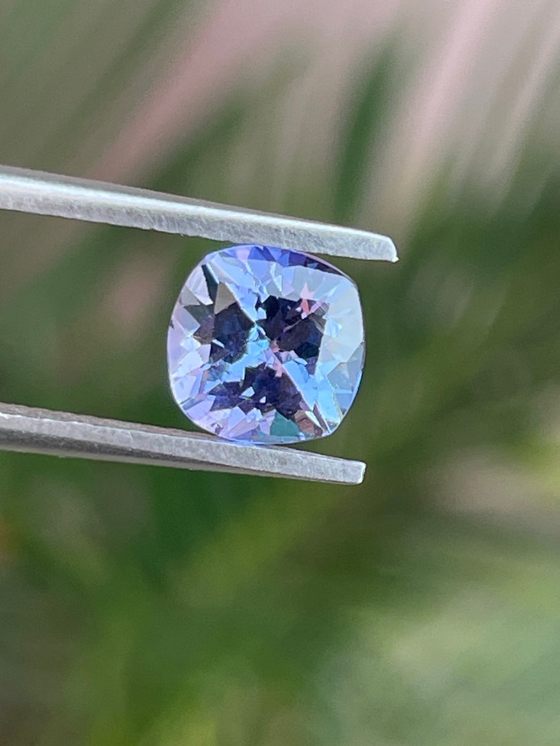 May include: A cushion-cut, purple gemstone with a light blue hue, held by tweezers against a blurred green background.