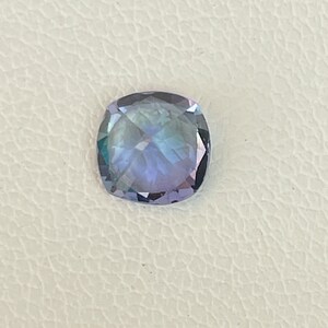 May include: A cushion-cut gemstone with a purple and blue hue. The gemstone is set against a white background.